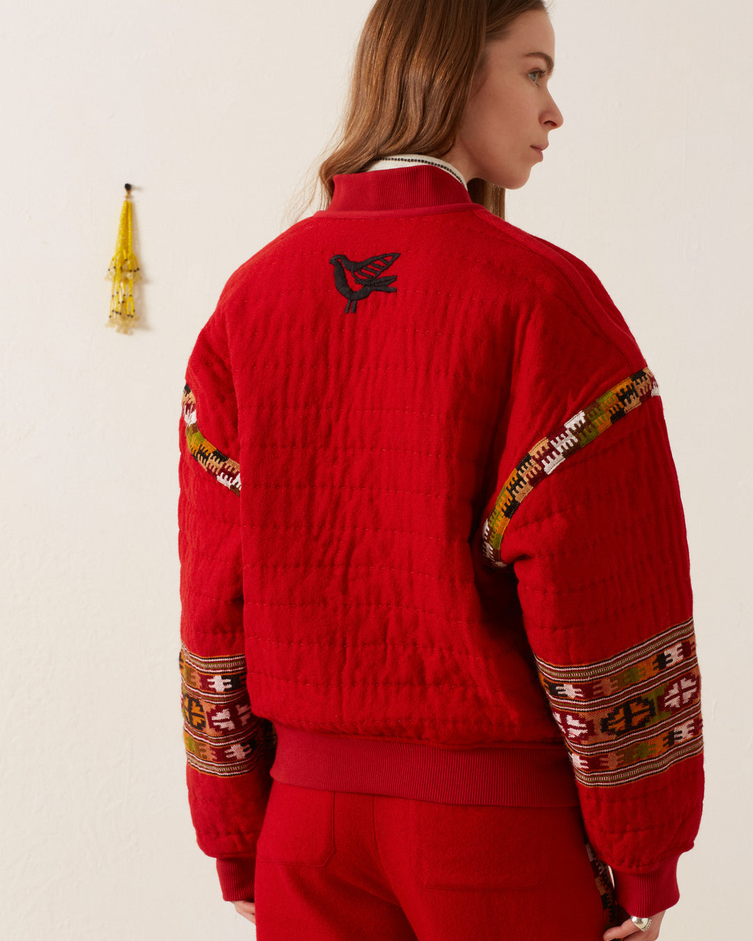 A person wearing the Sutton Scarlet Blanket Bomber, featuring a hand-quilted bird design on the back and decorative patterned sleeves, stands against a white wall with a yellow wall hanging.