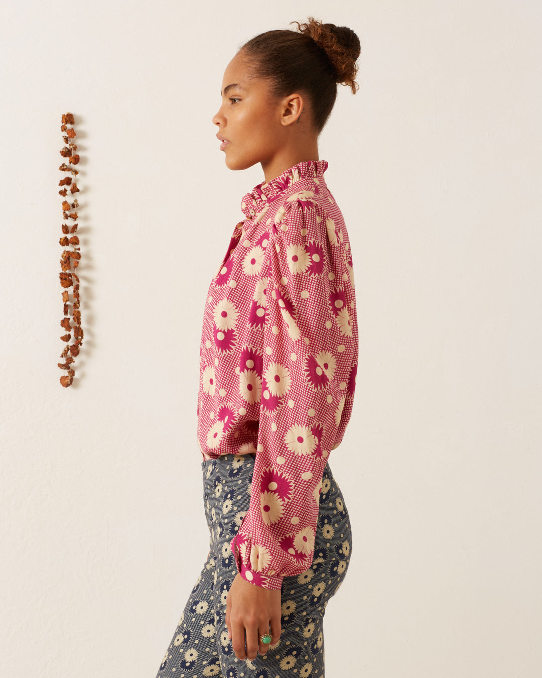 A person with a bun hairstyle stands in profile against a white wall. They are wearing the pink Annabel Berry Daisy Shirt, inspired by '70s vintage floral designs, along with blue pants adorned with white flowers. On the wall beside them is a vertical display of small brown objects.