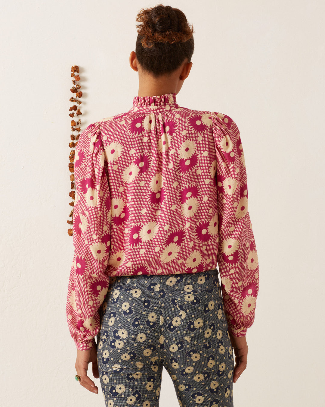 A person with short hair stands facing away, wearing the Annabel Berry Daisy Shirt, featuring a retro floral print in a pink blouse style, paired with blue pants showcasing a contrasting floral design. A vertical row of small decorative objects is mounted on the wall beside them, enhancing the '70s vintage-inspired vibe.