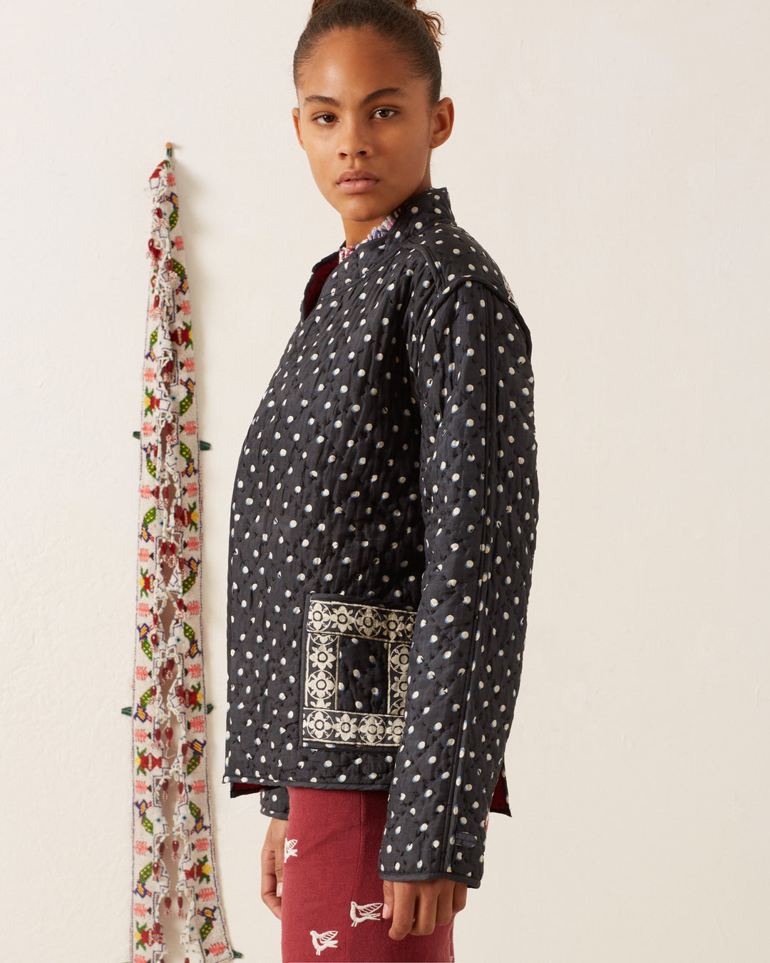 A person wearing a Tamil Raven Dot Silk Jacket with a patterned pocket stands against a white wall, next to a floral scarf hanging vertically.