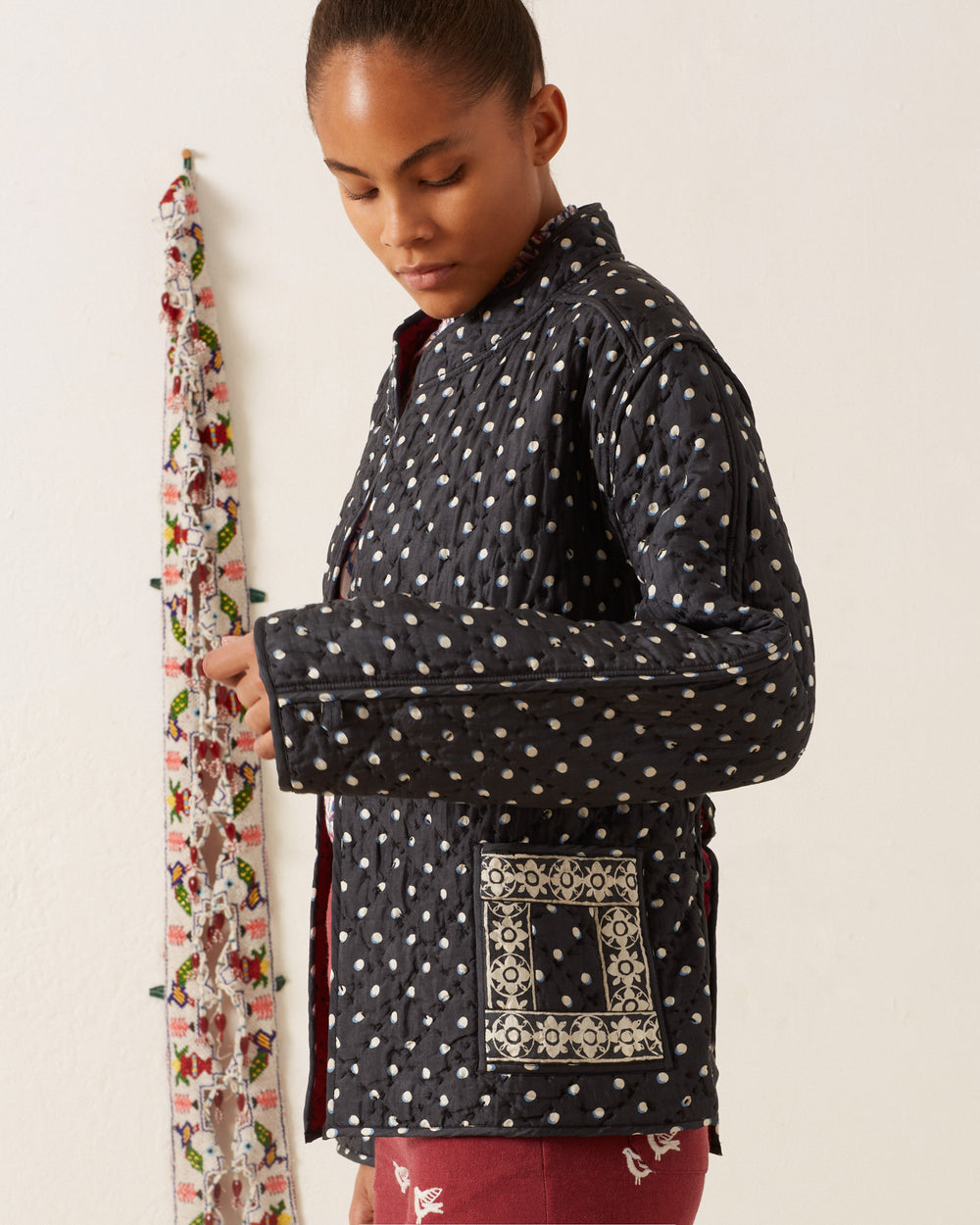 A person wearing the Tamil Raven Dot Silk Jacket, featuring a black hand-block printed design with a patterned pocket and tie-belt, stands sideways against a plain wall, as a colorful fabric strip hangs in the background.