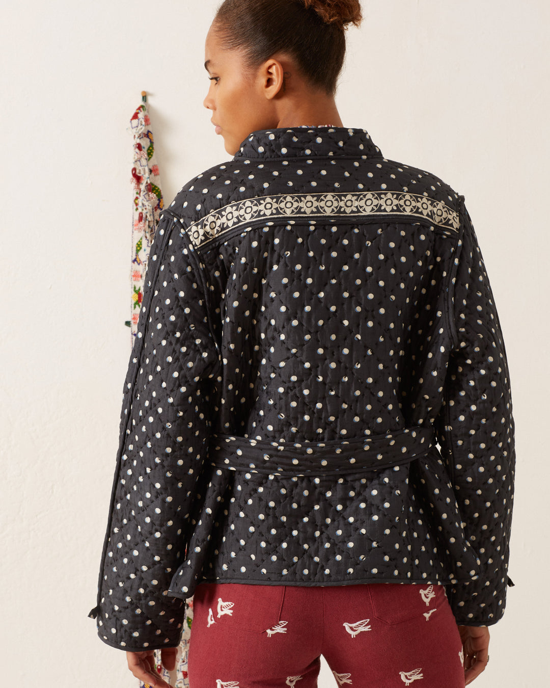 A person with their back turned is wearing the Tamil Raven Dot Silk Jacket, hand-block printed and adorned with decorative trim, along with maroon pants featuring a bird pattern.