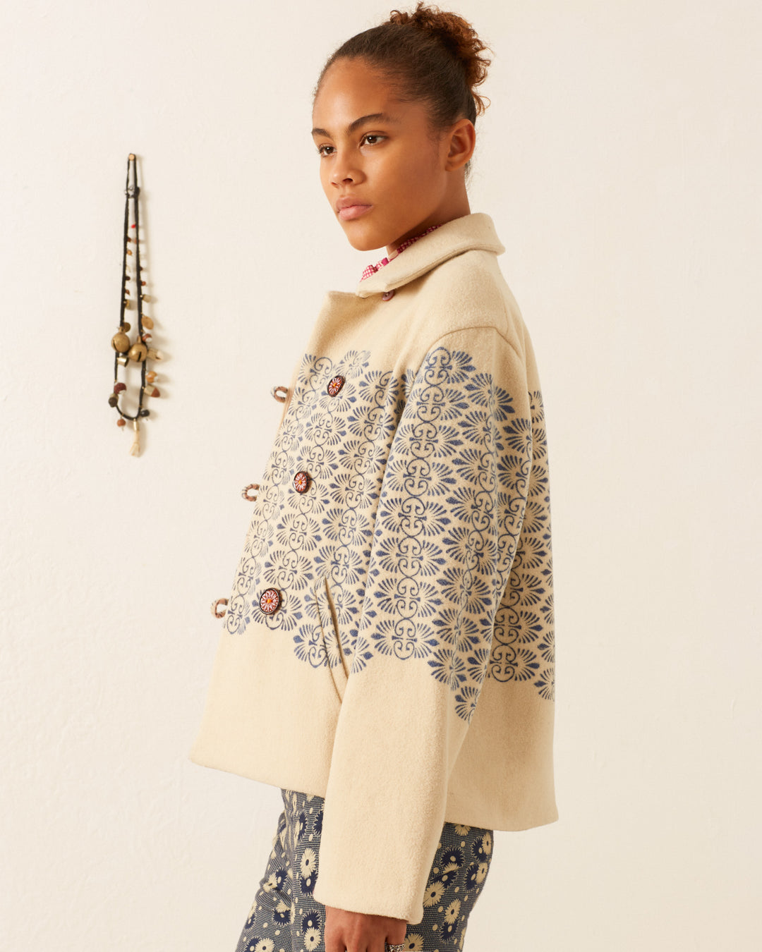 A person wearing a Western Lapis Trellis Jacket, featuring cream-colored fabric with blue floral designs, stands against a light-colored wall. Their dark hair is styled in a bun as they gaze to the side, while a decorative wall hanging adds charm to the scene.