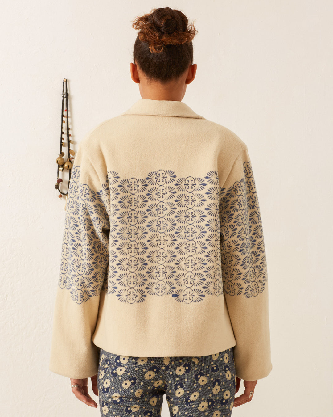 A person with short hair stands with their back to the camera, showcasing an ivory Western Lapis Trellis Jacket decorated with intricate blue patterns on the back and sleeves. The Western-inspired jacket pairs seamlessly with floral-patterned pants, while a beaded necklace is displayed on the wall.