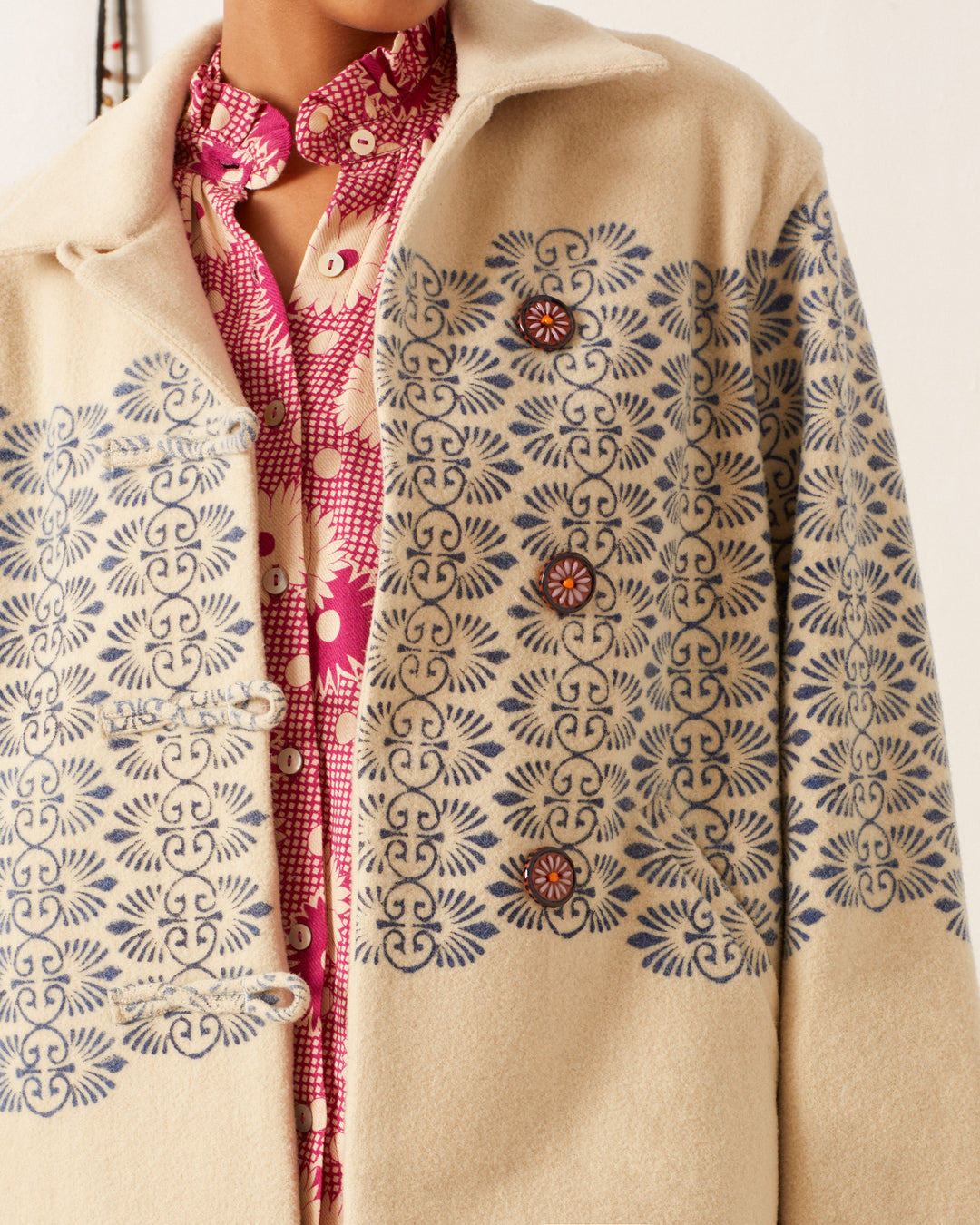 A close-up of someone wearing the Western Lapis Trellis Jacket, showcasing its elegant ivory fabric adorned with intricate blue floral patterns. This jacket is layered over a red and white floral patterned shirt and features unique buttons alongside detailed hand-block printed designs.