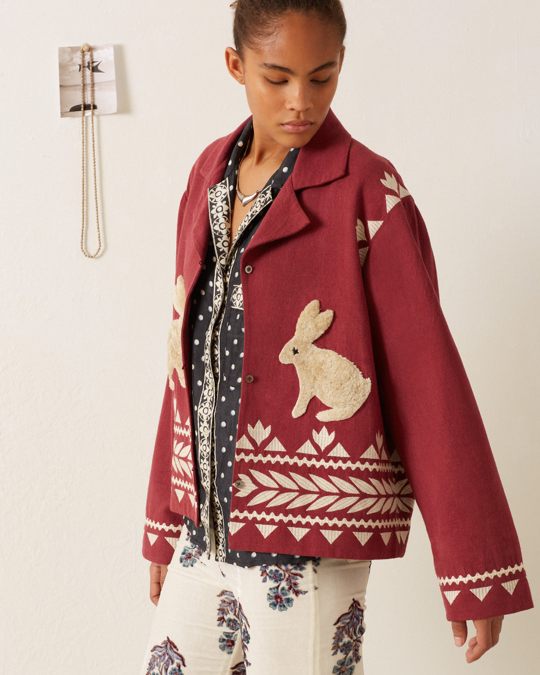 A person wears the Western Plum Hare Jacket, featuring maroon animal motifs with white rabbit and geometric patterns, as they stand near a wall. They pair it with a patterned shirt and floral pants, gazing slightly downward. In the background, a light wall with a hanging object completes the scene.