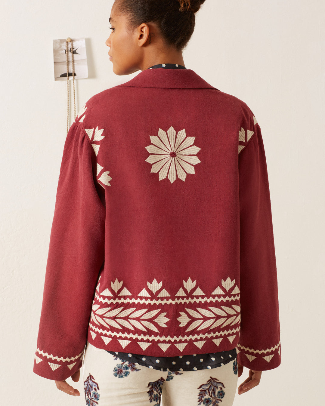 A person with short hair is turning away, wearing the Western Plum Hare Jacket, crafted from maroon silk and adorned with hand-embroidered white geometric patterns. The jacket showcases a prominent central star design, along with intricate decorative details along the hem and sleeves, all set against a plain white wall.