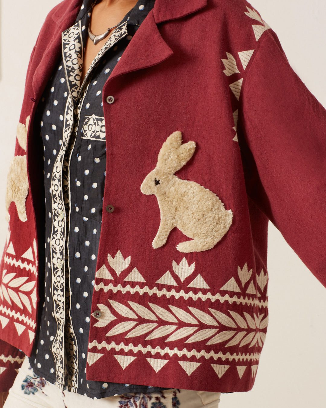 A person wearing a Western Plum Hare Jacket, showcasing white rabbit and geometric designs, layered over a black and white polka dot shirt. The lower part features clothing with floral patterns that highlight intricate hand embroidery.