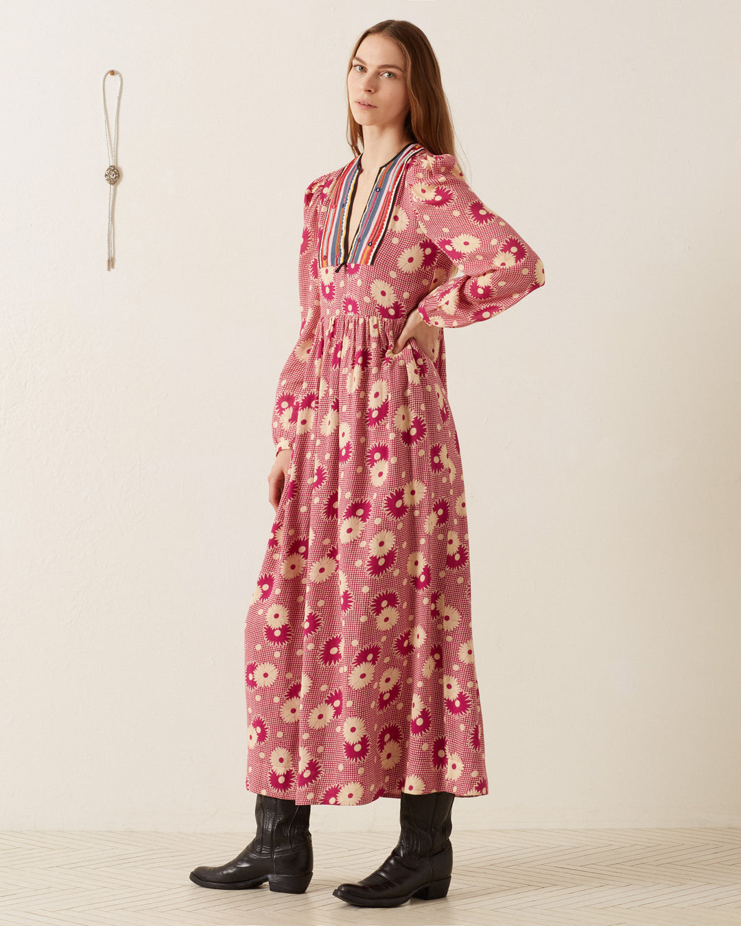 A woman in the Winifred Berry Daisy Dress, a long, retro floral prairie outfit with a colorful striped collar, stands on a tiled floor against a plain, light-colored wall. She is wearing black boots and looking toward the camera with her left hand on her hip, highlighting the romantic silhouette of the dress.