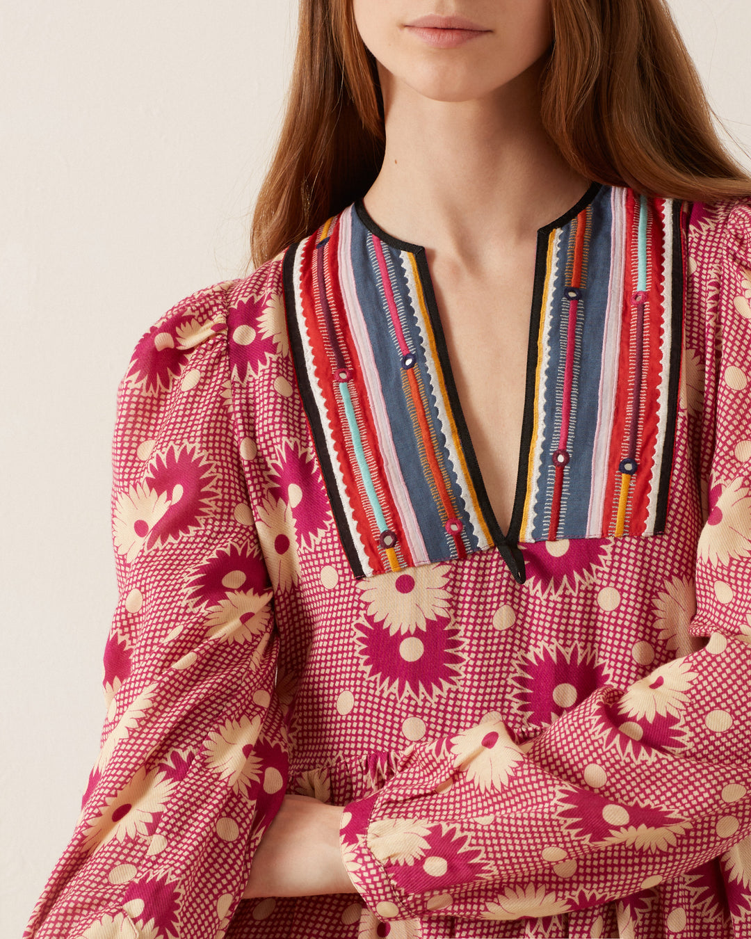 A person wears the Winifred Berry Daisy Dress, showcasing a vibrant retro floral prairie design with a red and pink pattern. It features a deep V-neck highlighted by multicolored stripes and embroidery. Made from a wool-cotton blend, this dress offers long sleeves and a textured design that enhances its romantic silhouette. The person has long, brown hair.