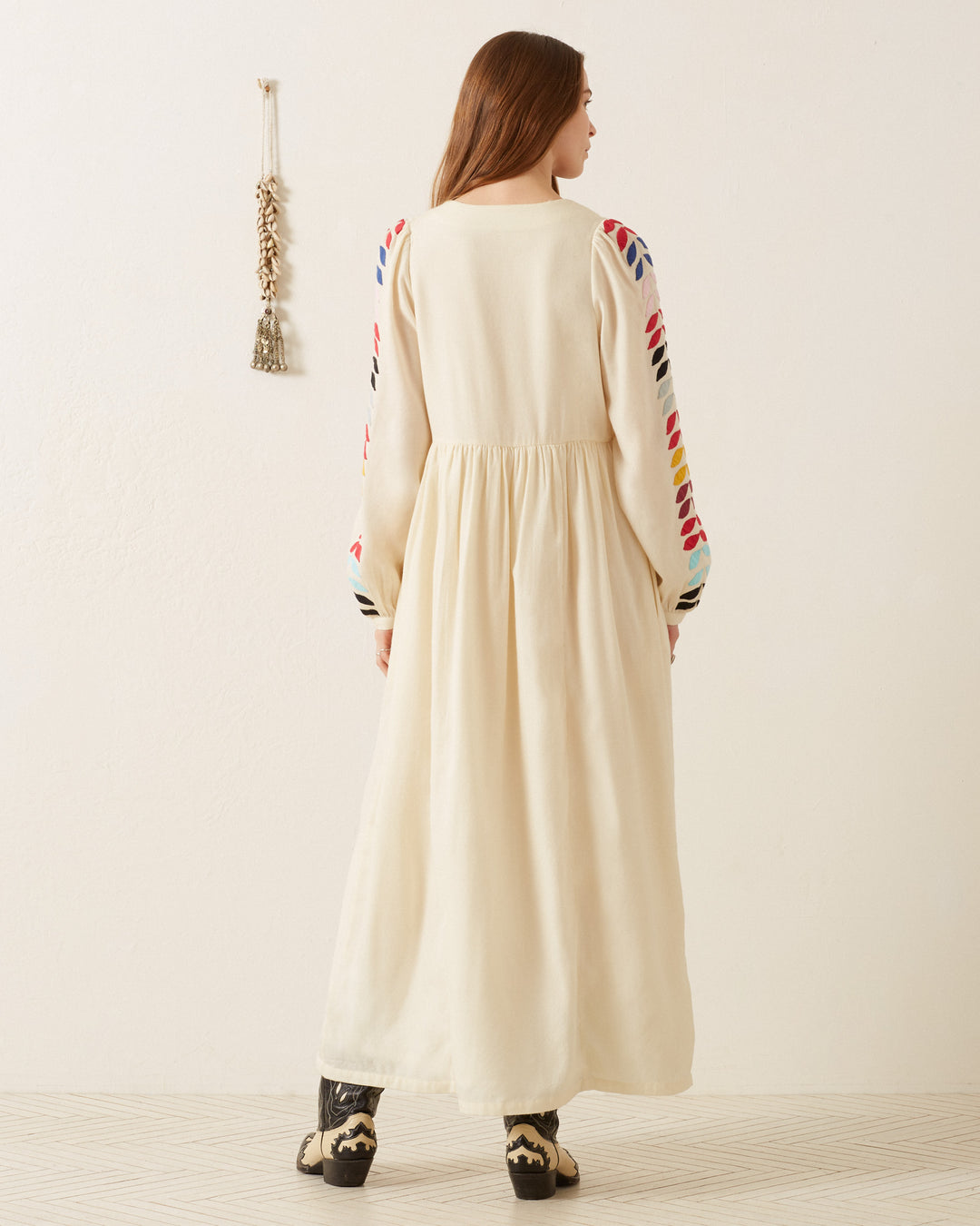 A person in an Imogen Ivory Prism Dress, featuring colorful embroidered sleeves and delicate floral appliqué, stands facing a white wall next to a hanging decorative item.