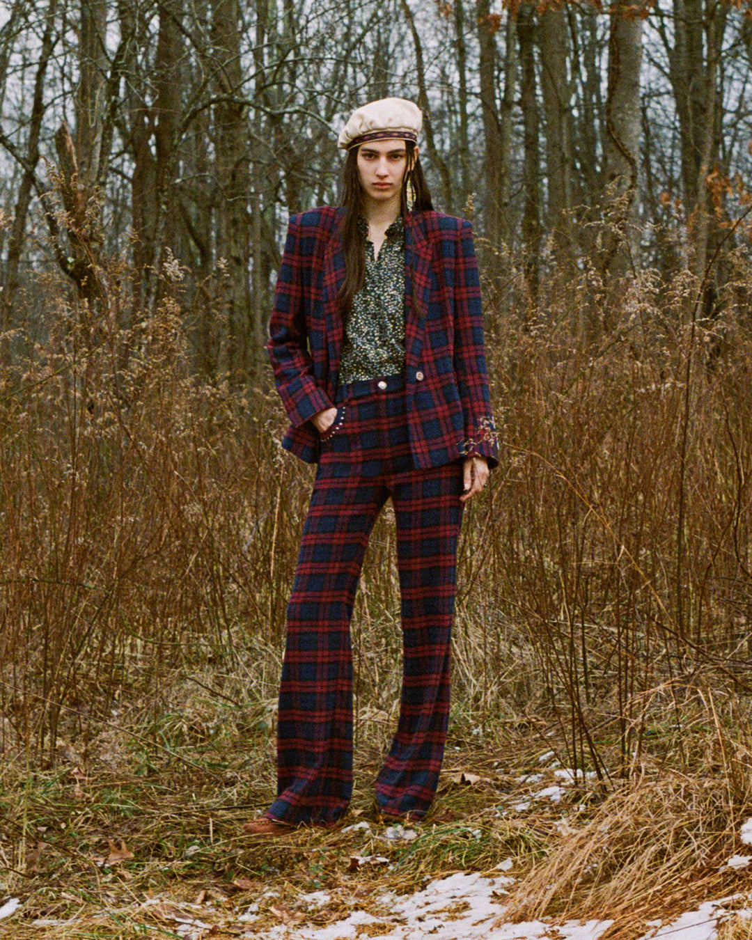 In a forest setting, an individual dons the Bunny Berry Plaid Pant paired with a floral shirt and hand-embellished details, complemented by a beige beret. The plaid pants harmonize with the rustic wintry landscape featuring lightly snow-dusted ground and bare trees.