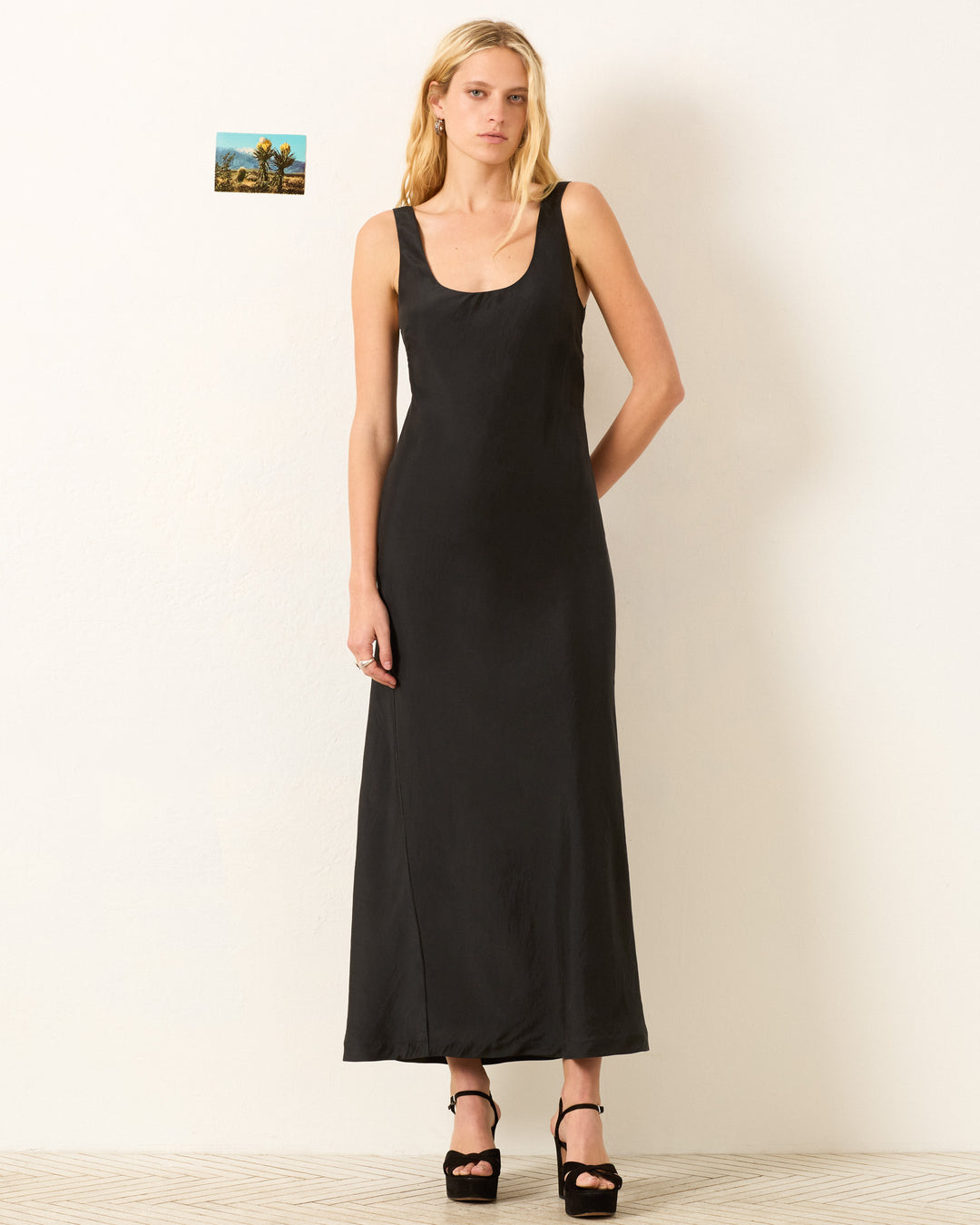 A woman stands against a white wall wearing the Adele Noir Silk Dress with black platform heels. A small photo is pinned to the wall behind her.