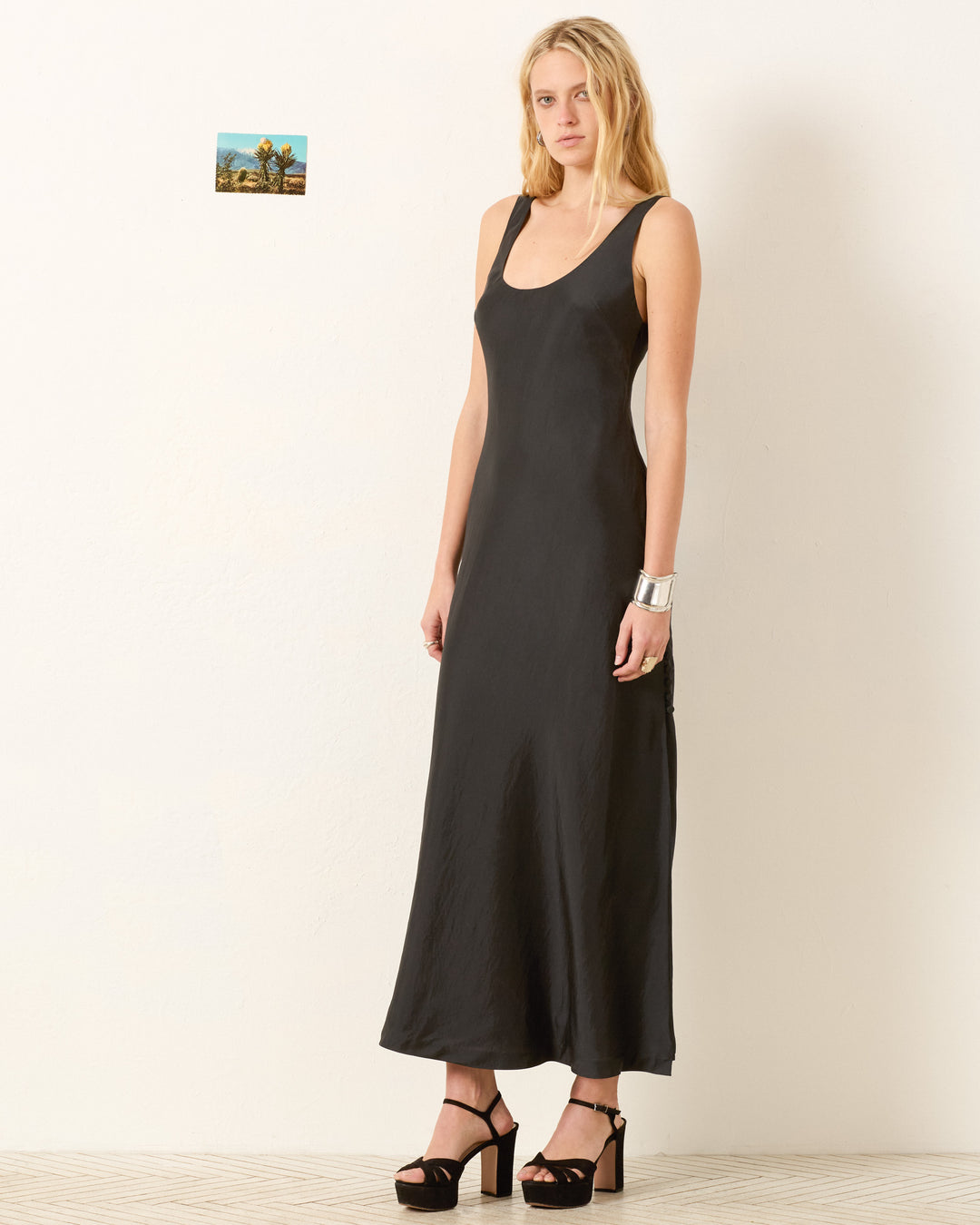 A woman stands indoors against a white wall wearing the Adele Noir Silk Dress and black platform heels, accessorized with a silver cuff bracelet. A small framed picture hangs on the wall behind her.