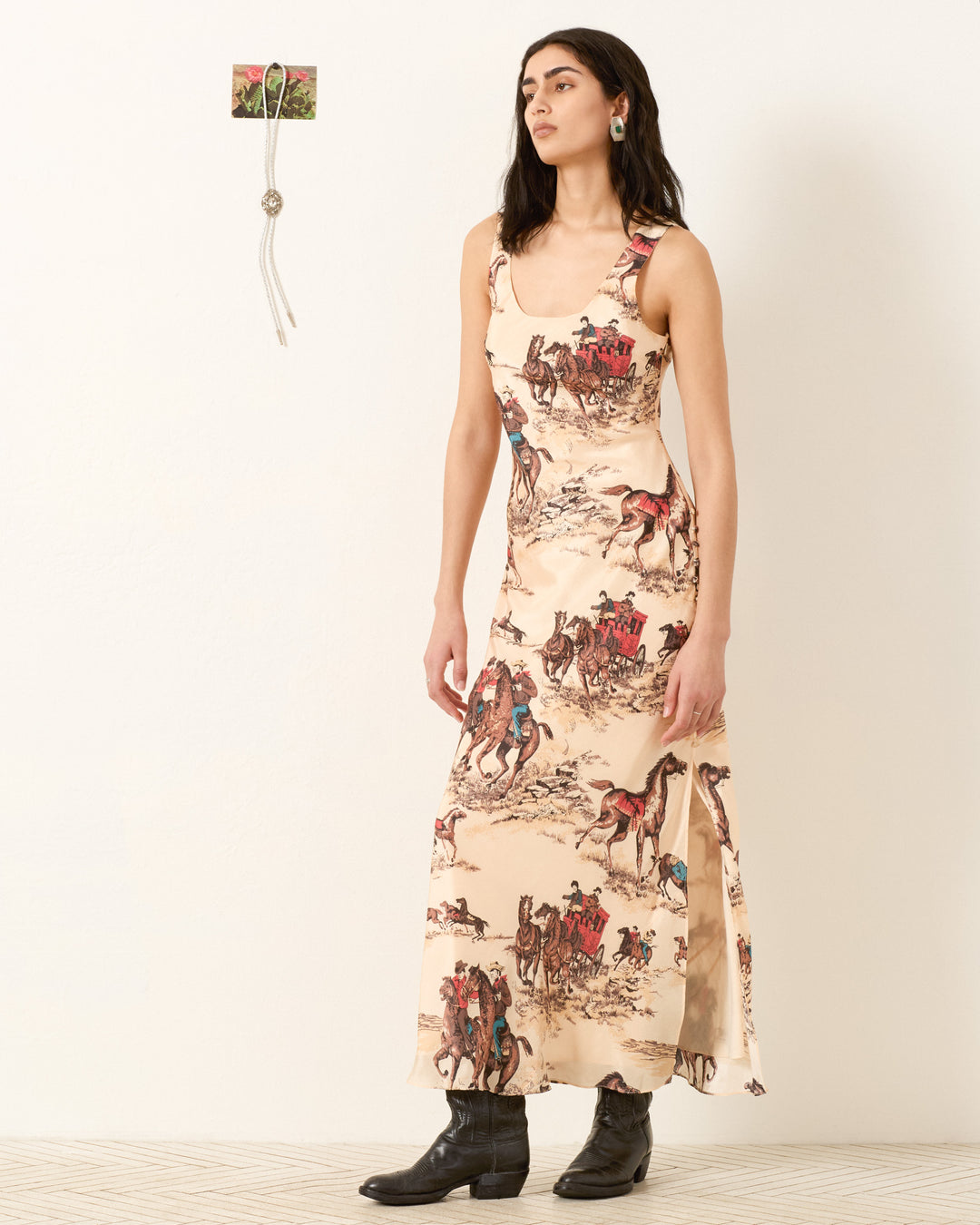Indoors, a woman wears the Adele Canyon Cowboy Silk Dress, a cream bias-cut maxi featuring a western horse and rider print, paired with black boots. A small decorative item hangs on the wall behind her.
