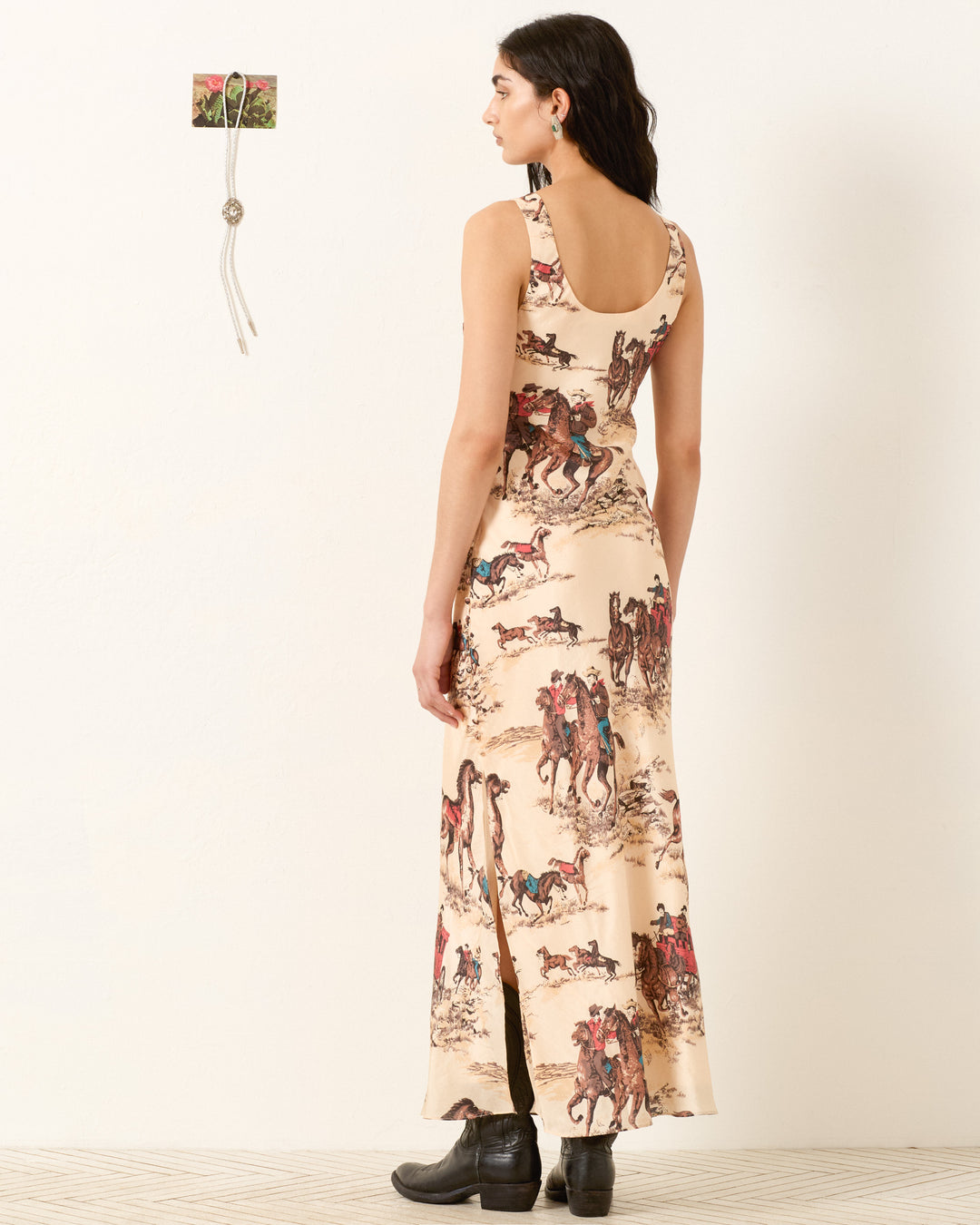 A woman stands indoors facing away, wearing the Adele Canyon Cowboy Silk Dress—a sleeveless, bias-cut maxi dress with a cowboy and horse print—paired with black boots.