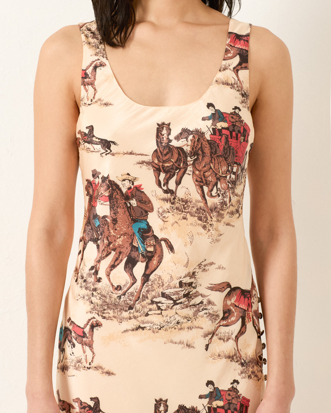 A woman wearing the Adele Canyon Cowboy Silk Dress, a sleeveless bias-cut maxi dress adorned with prints of cowboys on horseback and a horse-drawn carriage.