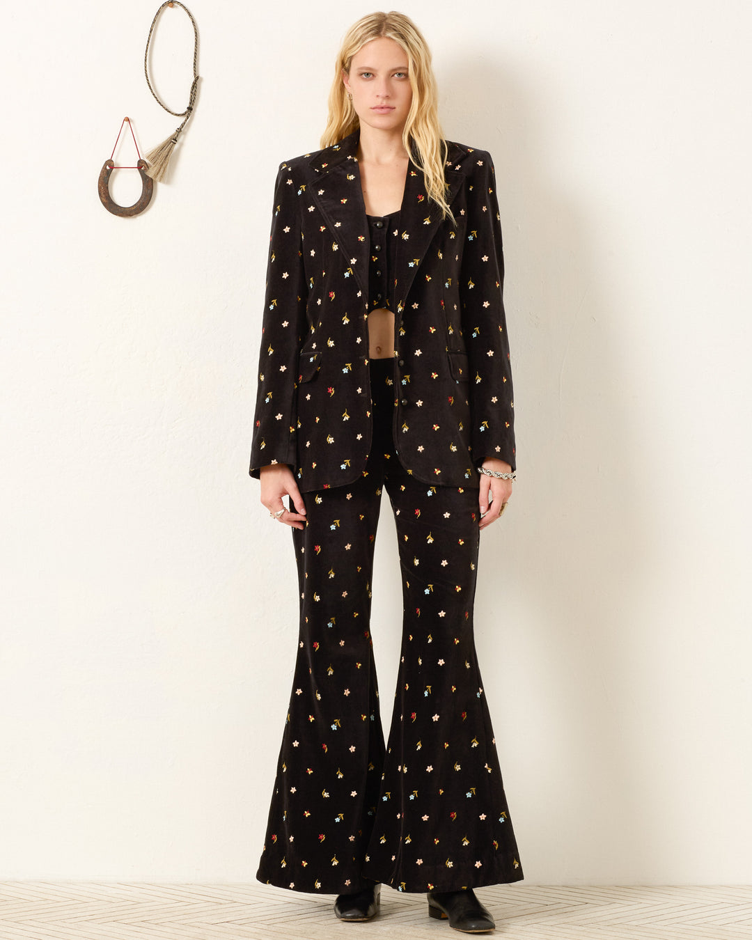 A woman stands against a white wall wearing the Afternoon Onyx Bouquet Blazer with floral embroidery, matching pants, a black top, and black boots. Decorative items hang on the wall beside her.
