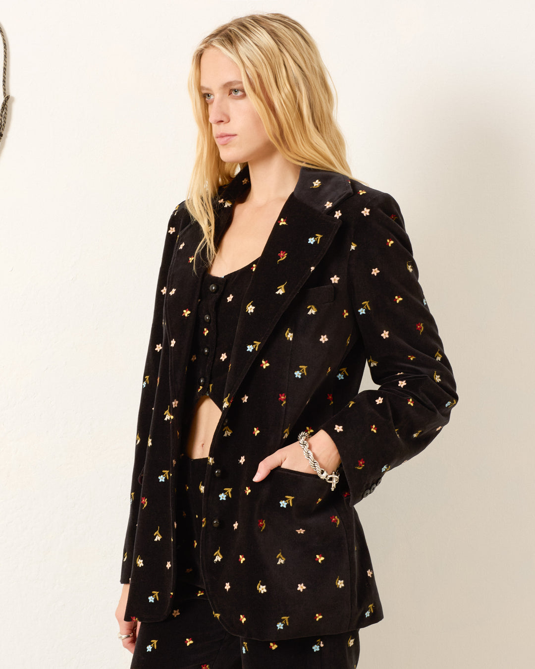 A woman with long blonde hair wears the Afternoon Onyx Bouquet Blazer, featuring multicolored floral embroidery, and matching pants while standing against a plain white wall.