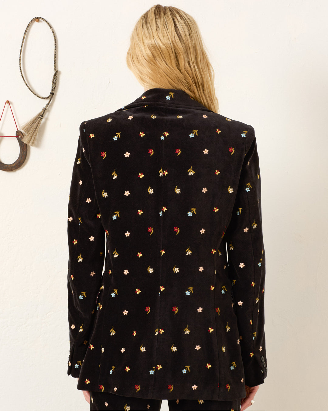 A person with long blonde hair seen from behind wears the Afternoon Onyx Bouquet Blazer, a black cotton-velvet jacket adorned with small multicolored floral embroidery. A decorative wall hanging appears to the left.