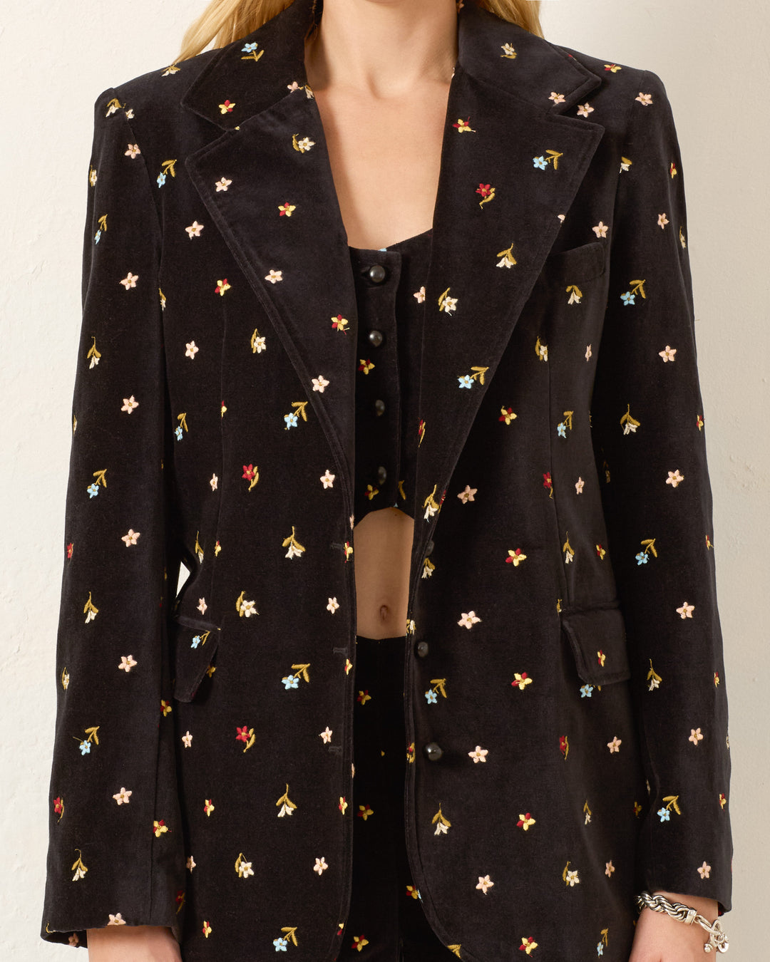A person wearing the Afternoon Onyx Bouquet Blazer, a black cotton-velvet blazer with multicolored floral embroidery, over a matching top and pants, shown from shoulders to hips.