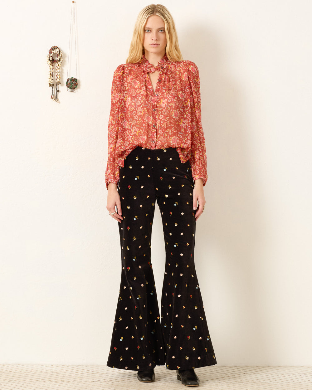A woman stands against a white wall wearing the Annabel Sunset Ember Shirt with puff shoulders and black flared pants featuring a small floral print. She completes her look with black shoes, decorative wall hangings visible behind her.