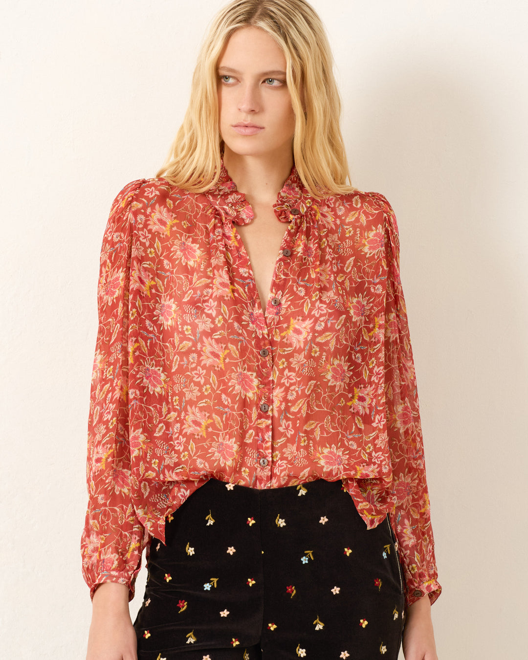 A woman wears the Annabel Sunset Ember Shirt, a sheer red floral viscose-chiffon top with a ruffled collar and puff shoulders, paired with black pants featuring small embroidered flowers, standing against a plain light background.