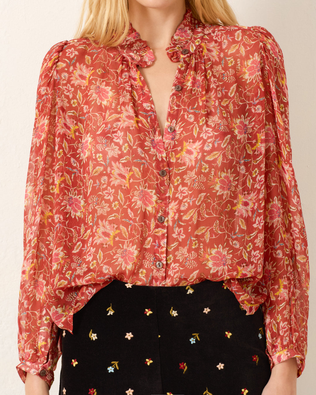 A woman wears the Annabel Sunset Ember Shirt, a long-sleeve red floral viscose-chiffon top with a ruffled collar and puff shoulders, paired with a black skirt featuring small embroidered flowers.