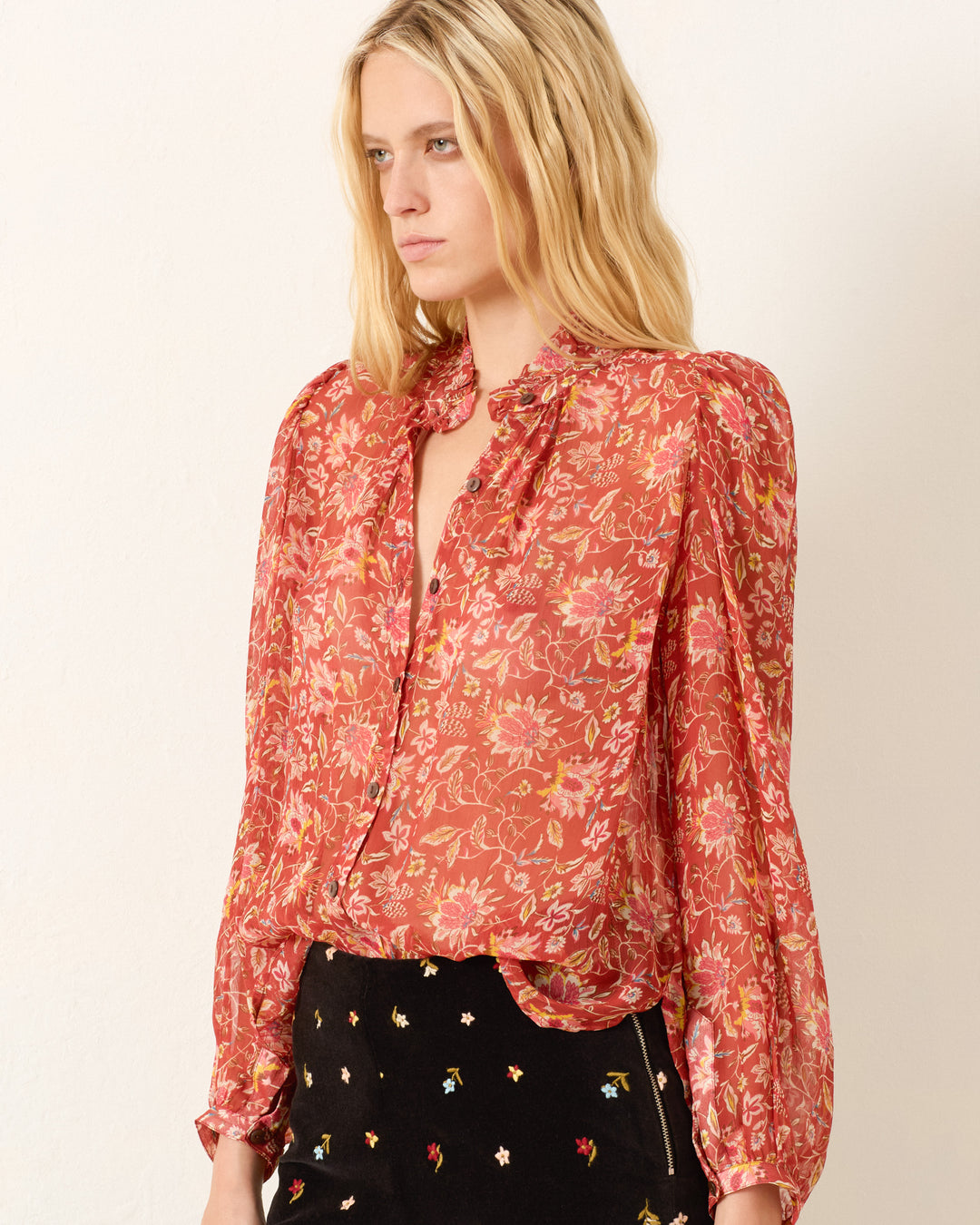 A woman stands against a plain background wearing the Annabel Sunset Ember Shirt—a red floral viscose-chiffon top with puff shoulders and a ruffled collar—paired with black pants featuring small embroidered flowers.