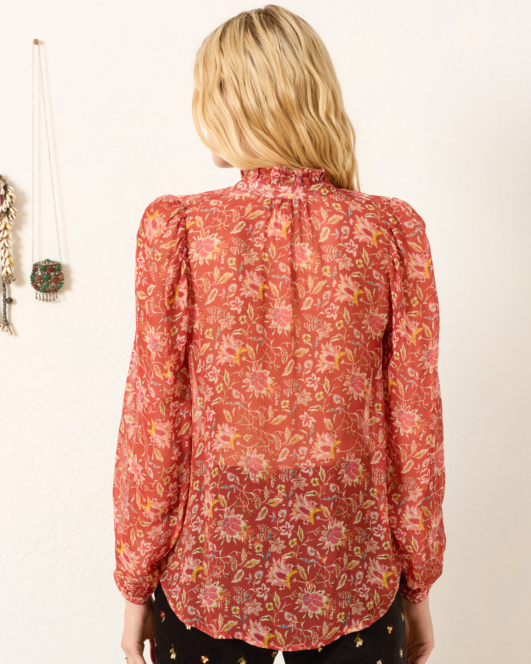 A person with long blonde hair is seen from behind, wearing the Annabel Sunset Ember Shirt—a red floral-patterned viscose-chiffon top with puff shoulders and a high collar. Decorative items hang on the white wall.