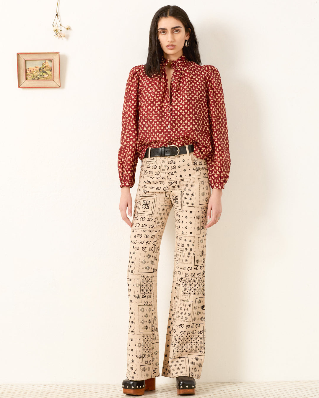 A person stands against a white wall wearing the Annabel Mahogany Bhutti Shirt, beige hand-block printed wide-leg pants, a black belt, and platform shoes. Nearby, a small framed picture and dried flower hang on the wall.