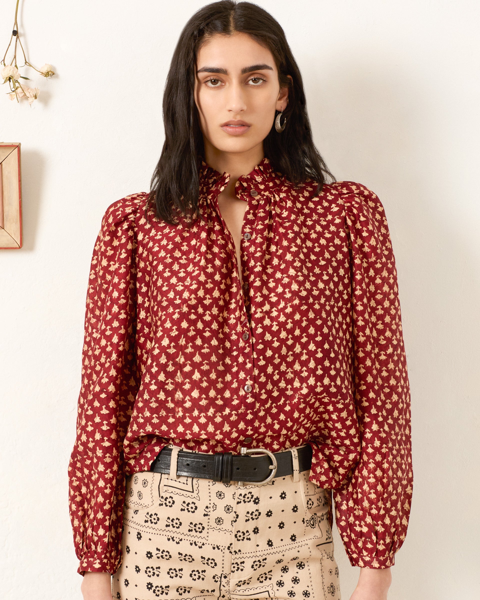 A woman with long dark hair wears the Annabel Mahogany Bhutti Shirt, a vintage-inspired red patterned blouse with puffed sleeves, paired with beige high-waisted pants and a black belt, standing against a plain white wall.