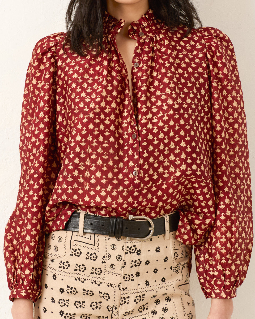 A woman wears the Annabel Mahogany Bhutti Shirt, a vintage-inspired, long-sleeve red blouse with a small cream floral print, paired with high-waisted beige pants featuring a black floral pattern and a black belt.
