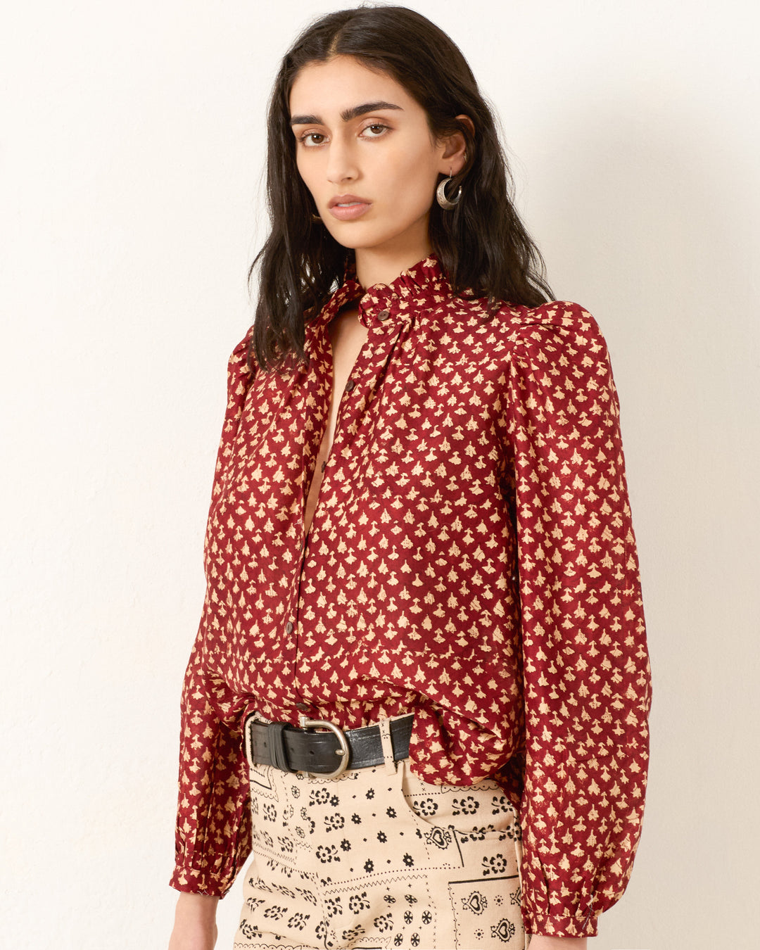 A woman wears the Annabel Mahogany Bhutti Shirt, a vintage-inspired red blouse with a yellow floral pattern, paired with beige high-waisted pants and a black belt, standing against a plain white background.