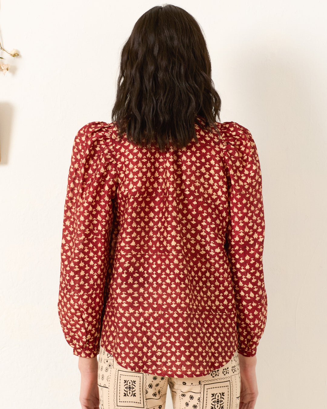 Seen from behind, a person with wavy dark hair wears the Annabel Mahogany Bhutti Shirt—a vintage-inspired red patterned blouse with long sleeves—paired with cream patterned pants against a plain light background.