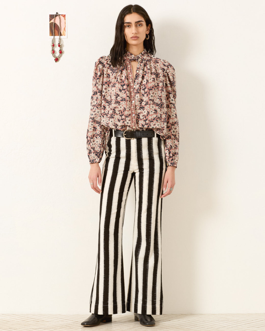 A woman stands against a white wall wearing the Annabel Sepia Floral Shirt, black and white vertically striped flared pants, a black belt, and black shoes. A small framed picture hangs above her left shoulder.
