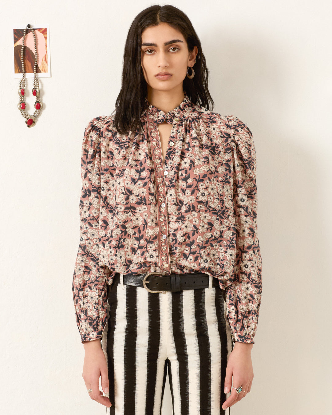 A woman stands against a white wall wearing the Annabel Sepia Floral Shirt with a tie-neck detail, paired with black-and-white striped pants, a belt, and jewelry.