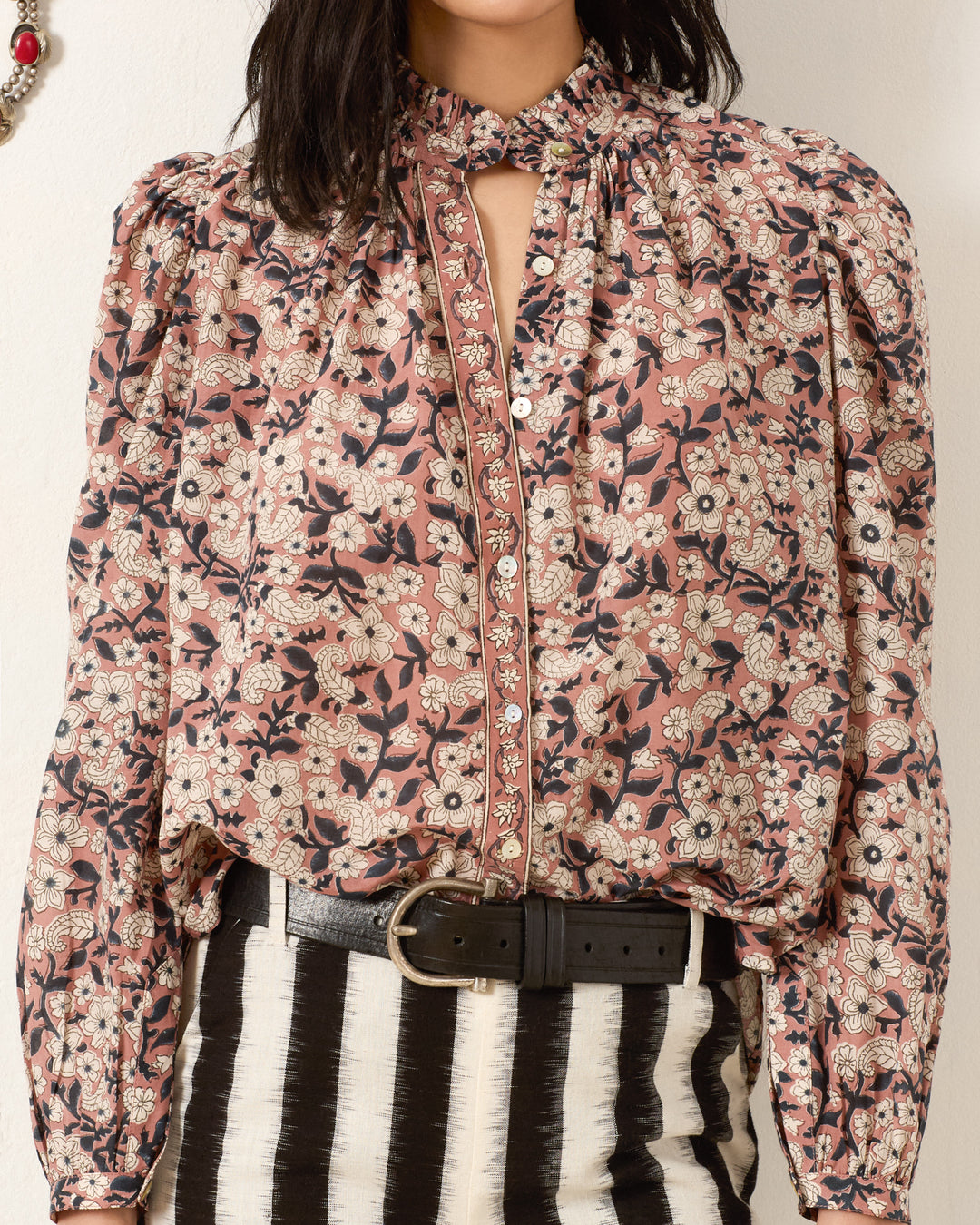 A person wears the Annabel Sepia Floral Shirt, featuring a cotton-silk blend with hand-block printed florals, ruffled collar, and button details, paired with high-waisted black and white striped pants and a black belt.