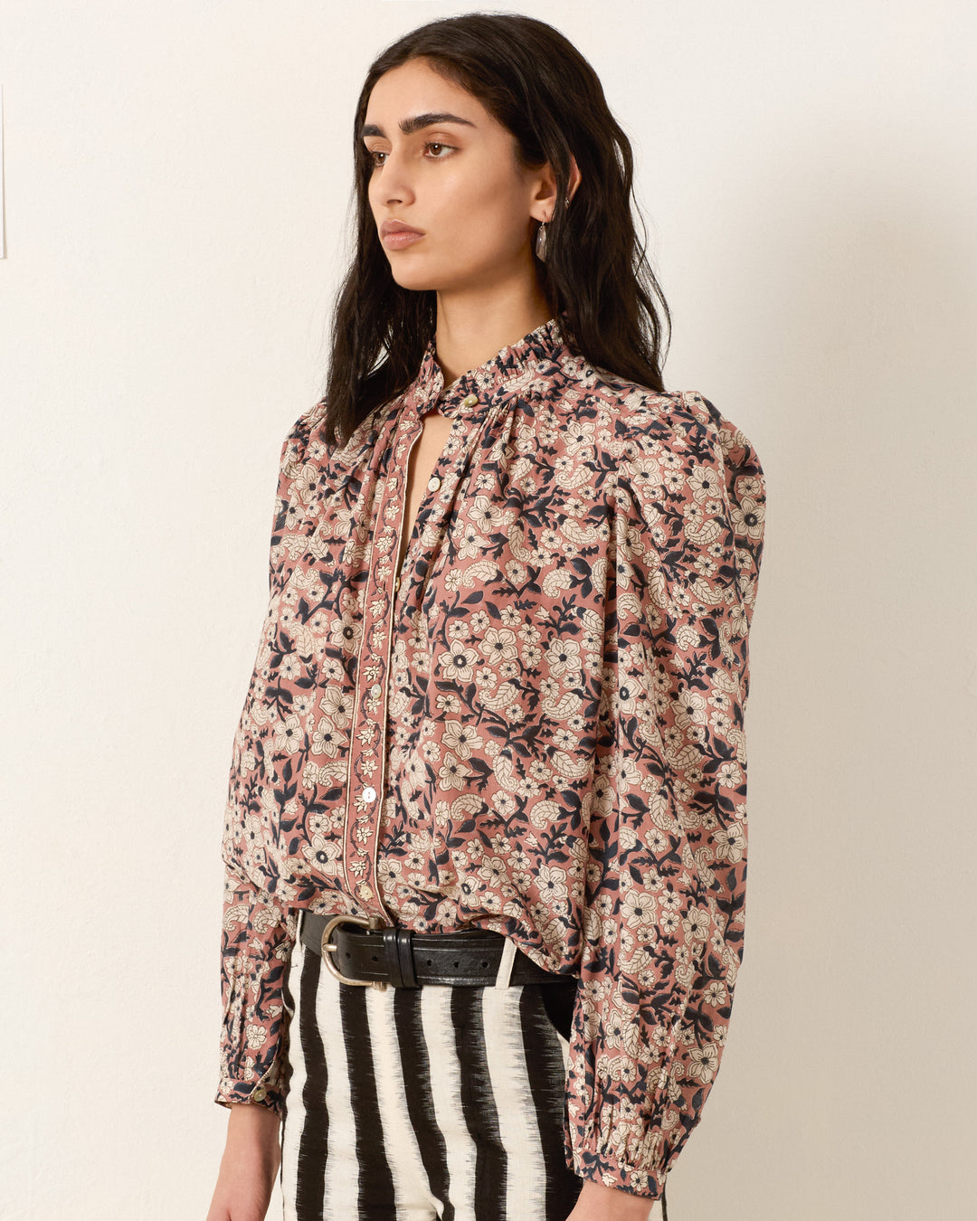 A woman stands against a plain wall wearing the Annabel Sepia Floral Shirt, a cotton-silk blend with hand-block printed florals, high neck, and puffed sleeves, paired with black and white striped pants and a black belt.