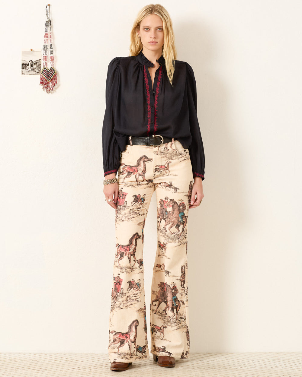 A woman stands against a white wall wearing the Annabel Raven Wing Shirt and high-waisted cream-colored pants with a horse and rider print.