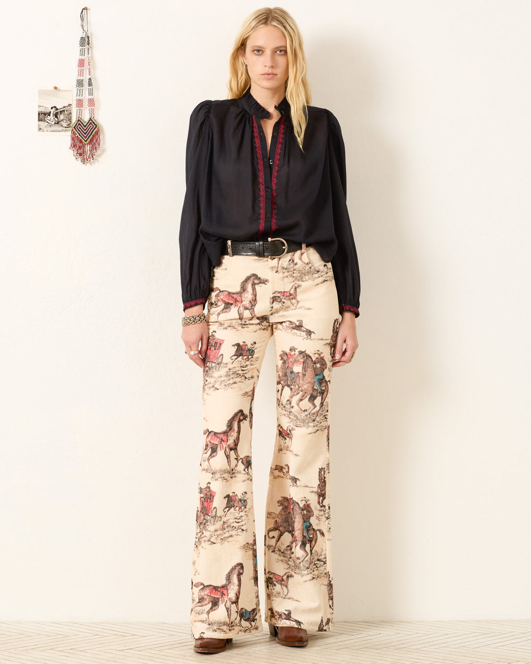 A woman stands against a white wall wearing the Annabel Raven Wing Shirt and high-waisted cream-colored pants with a horse and rider print.