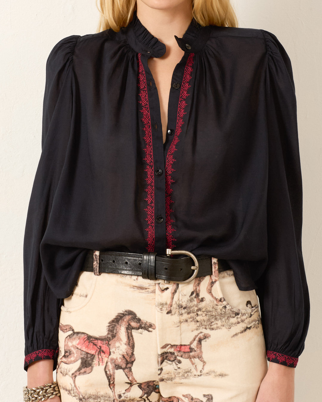 A person wears the Annabel Raven Wing Shirt, a black long-sleeve hand-embroidered blouse with red trim, paired with cream horse-print pants and a black belt.