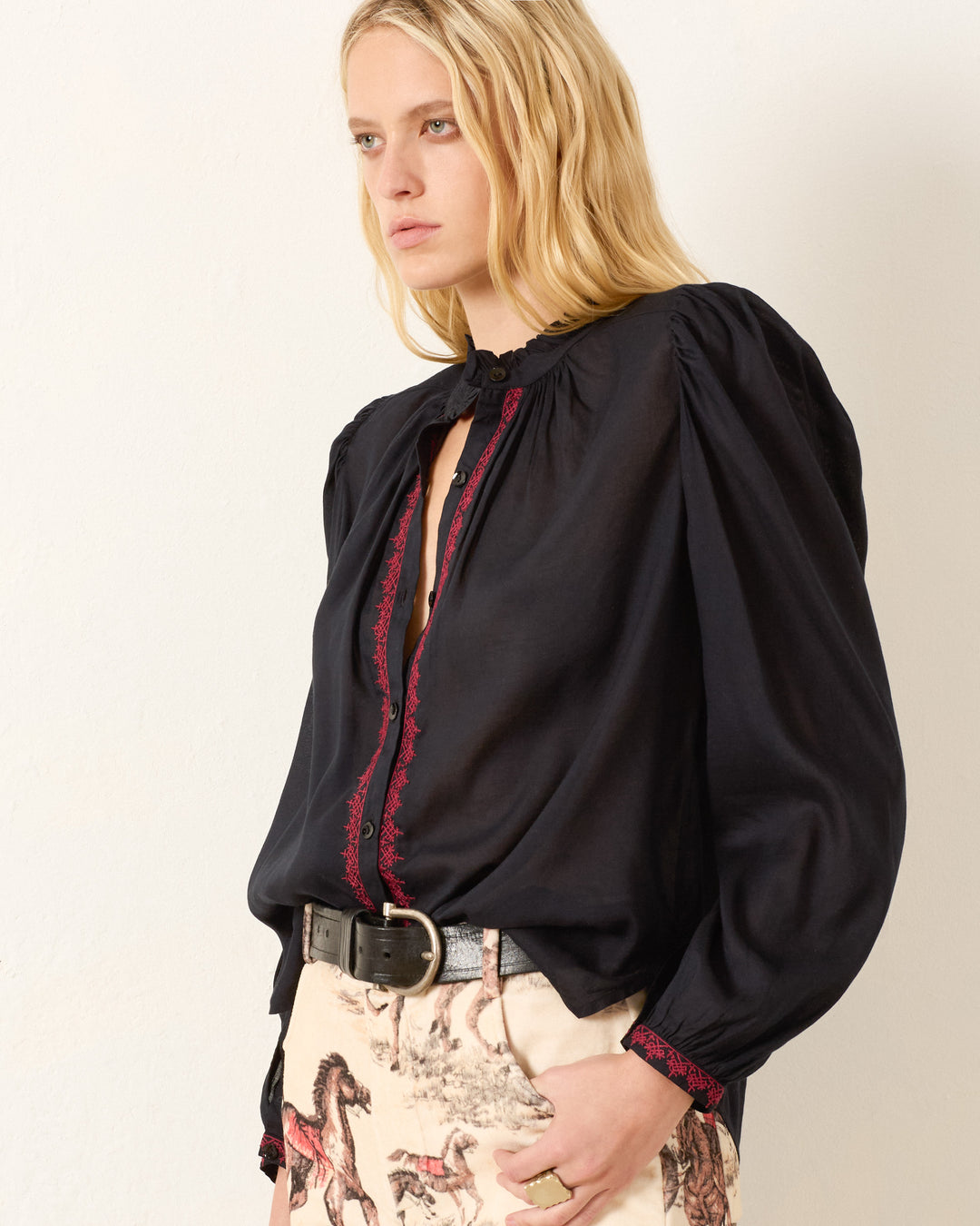 A woman with long blonde hair wears the Annabel Raven Wing Shirt, a vintage-inspired black blouse with red trim and hand embroidery, paired with cream horse-print pants, standing against a plain light background.