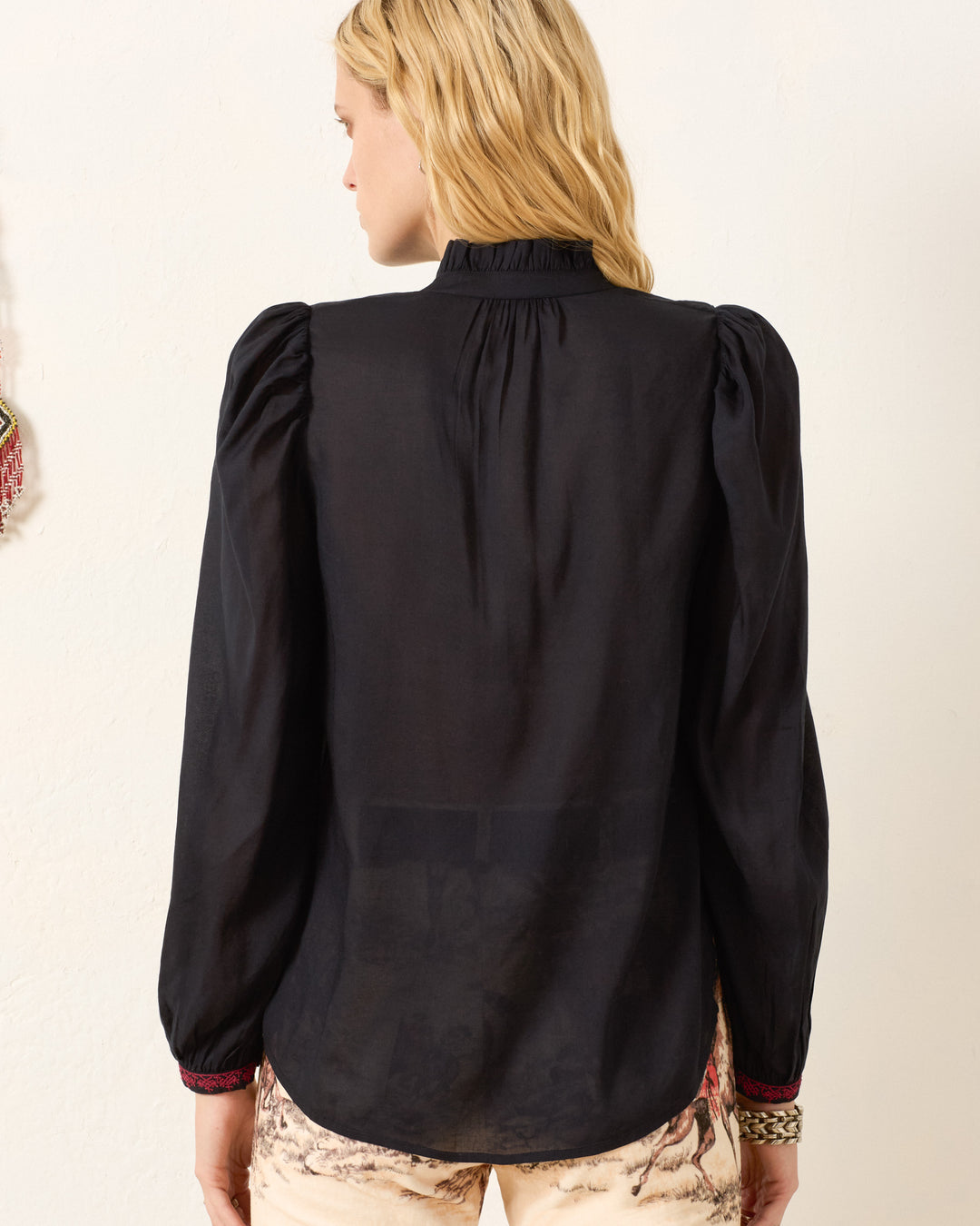 A person with wavy blonde hair is shown from the back, wearing the Annabel Raven Wing Shirt—a long-sleeve black vintage-inspired top with gathered shoulders—and light-colored pants.