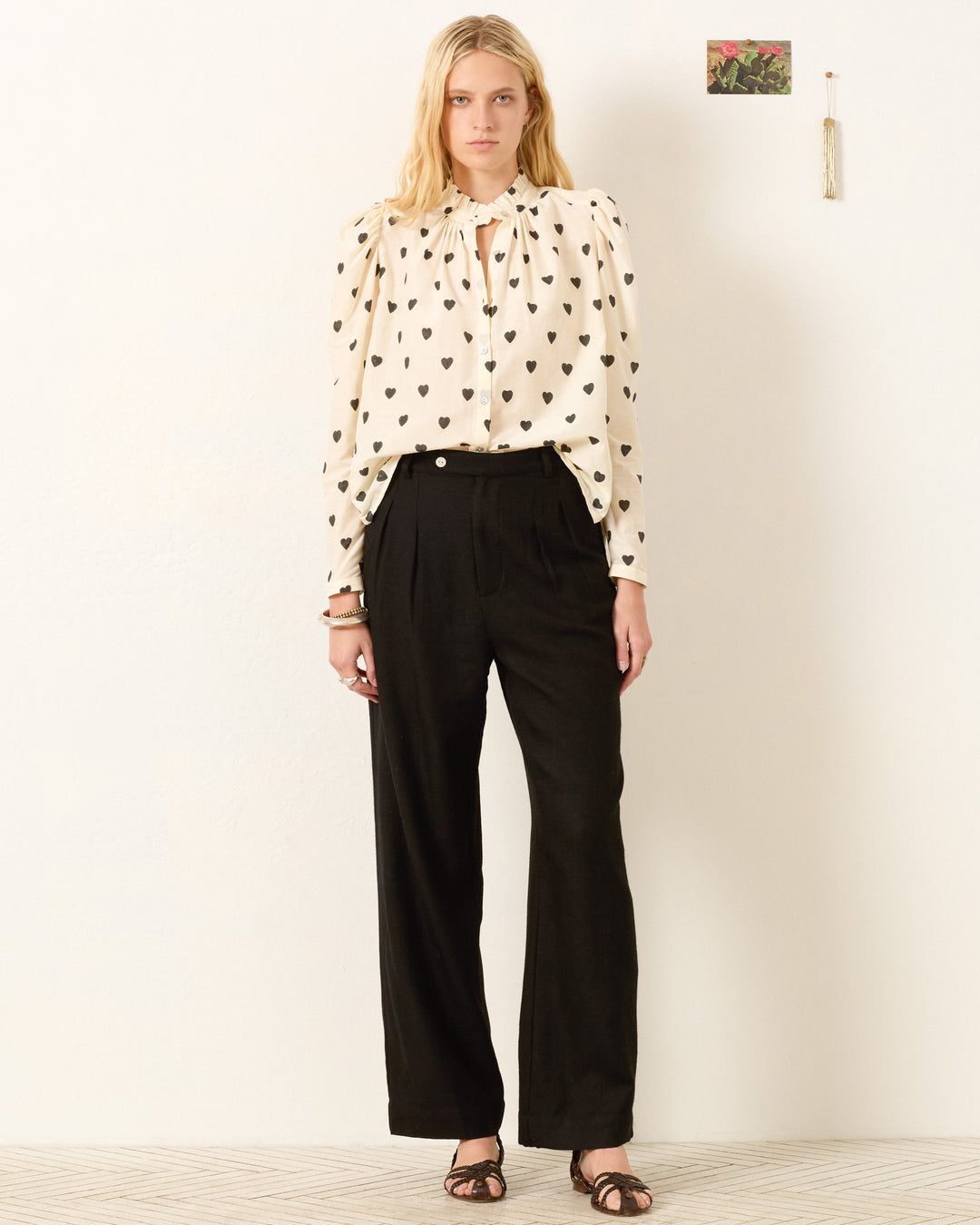 A woman stands against a plain wall wearing the Artemis Onyx Heart Shirt, black high-waisted trousers, and black sandals.