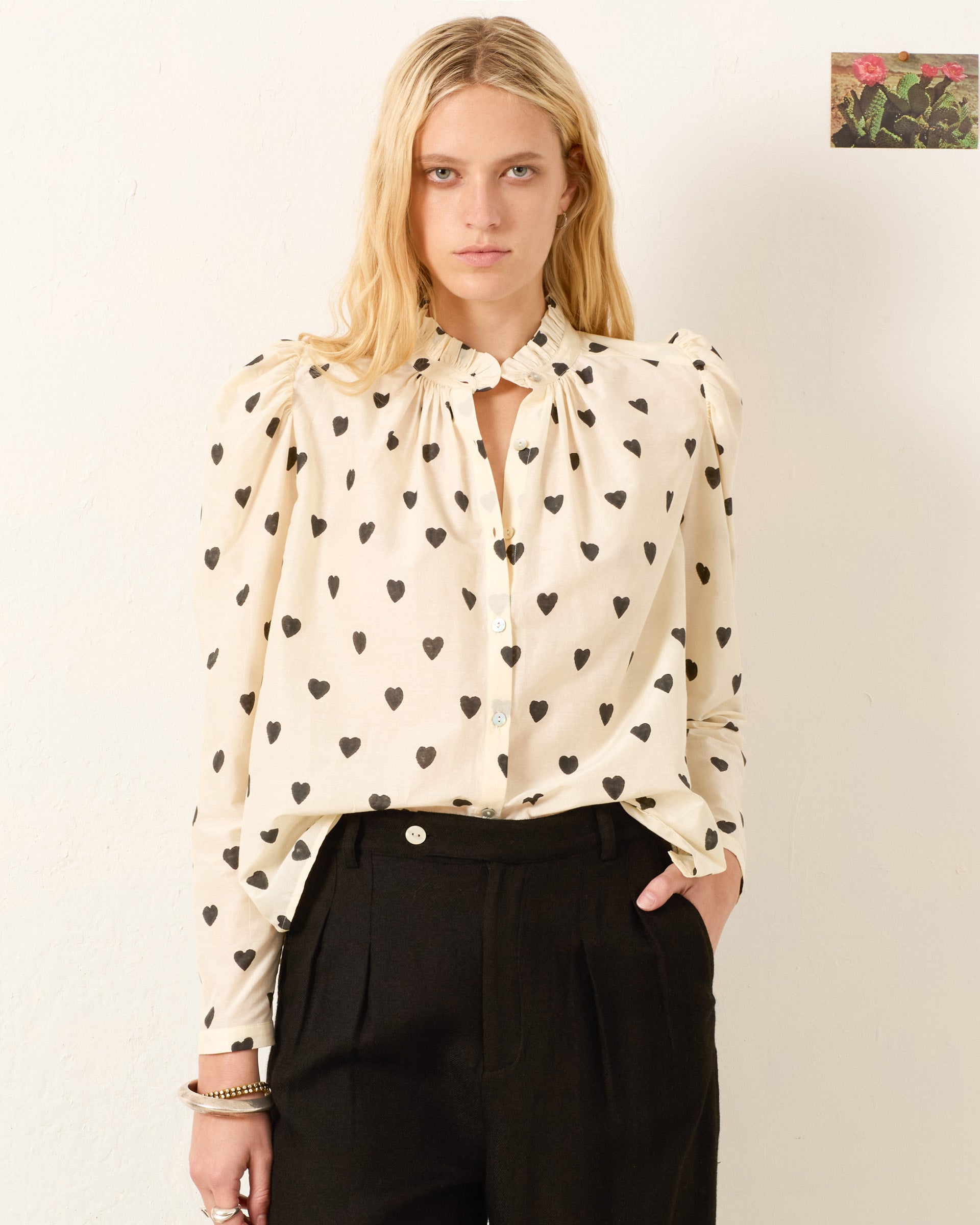 A woman with long blonde hair wears the Artemis Onyx Heart Shirt, featuring black heart patterns, paired with black trousers, standing against a plain, light-colored wall.