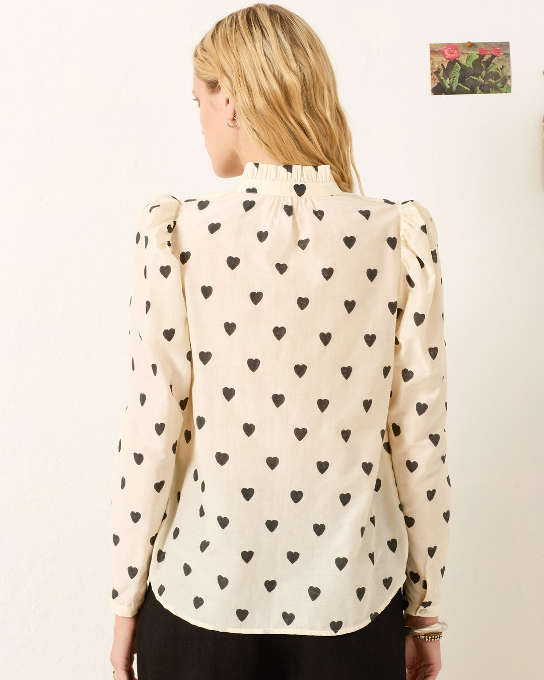 A person with long blonde hair is seen from behind wearing the Artemis Onyx Heart Shirt, a cream hand-block-printed blouse with a black heart pattern and ruffled collar; a small flower photo is on the wall in the background.