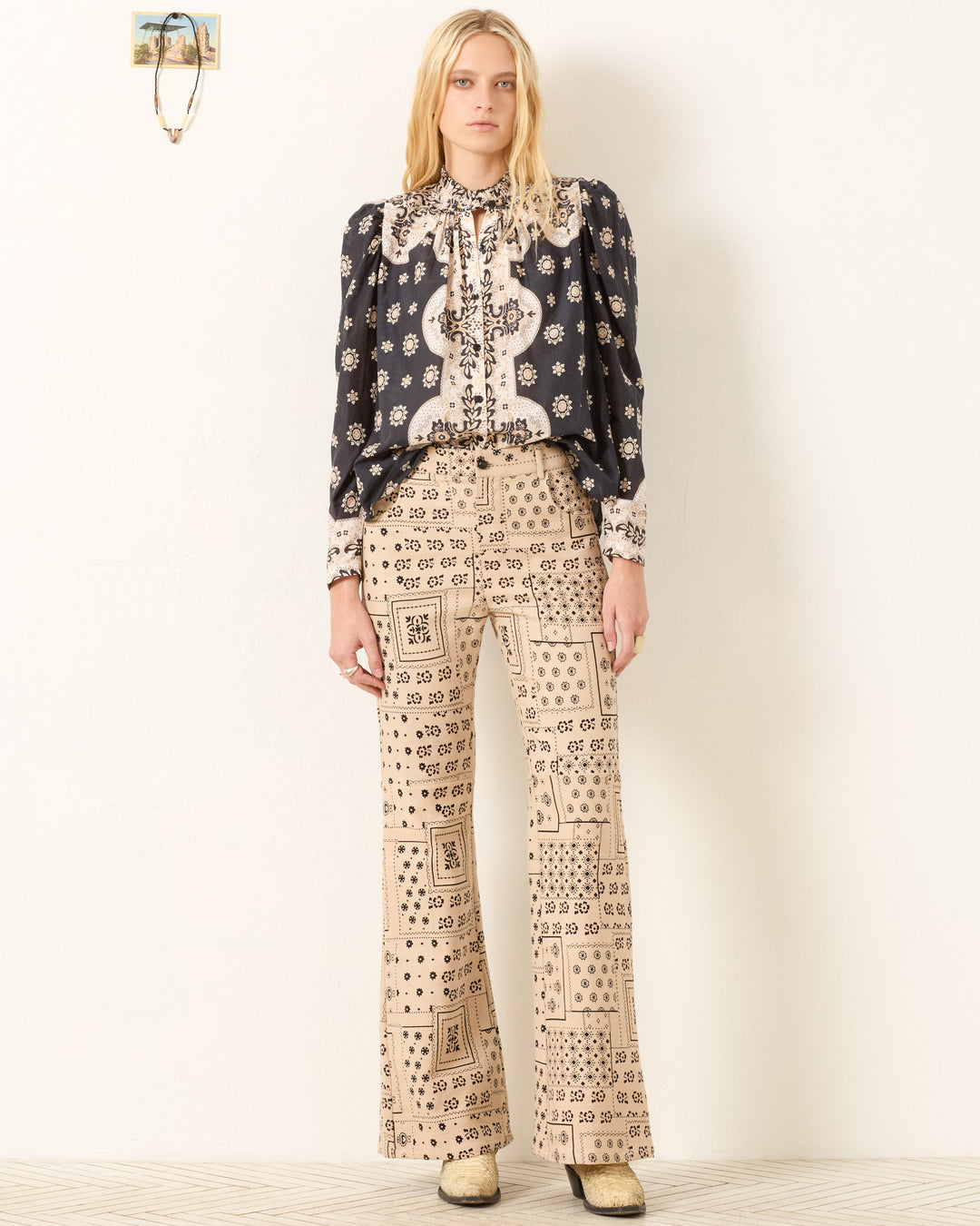 A person with long, straight hair and a neutral expression stands against a plain wall wearing the Artemis Bronco Bandana Shirt with puff sleeves and beige high-waisted flared pants featuring geometric patterns.