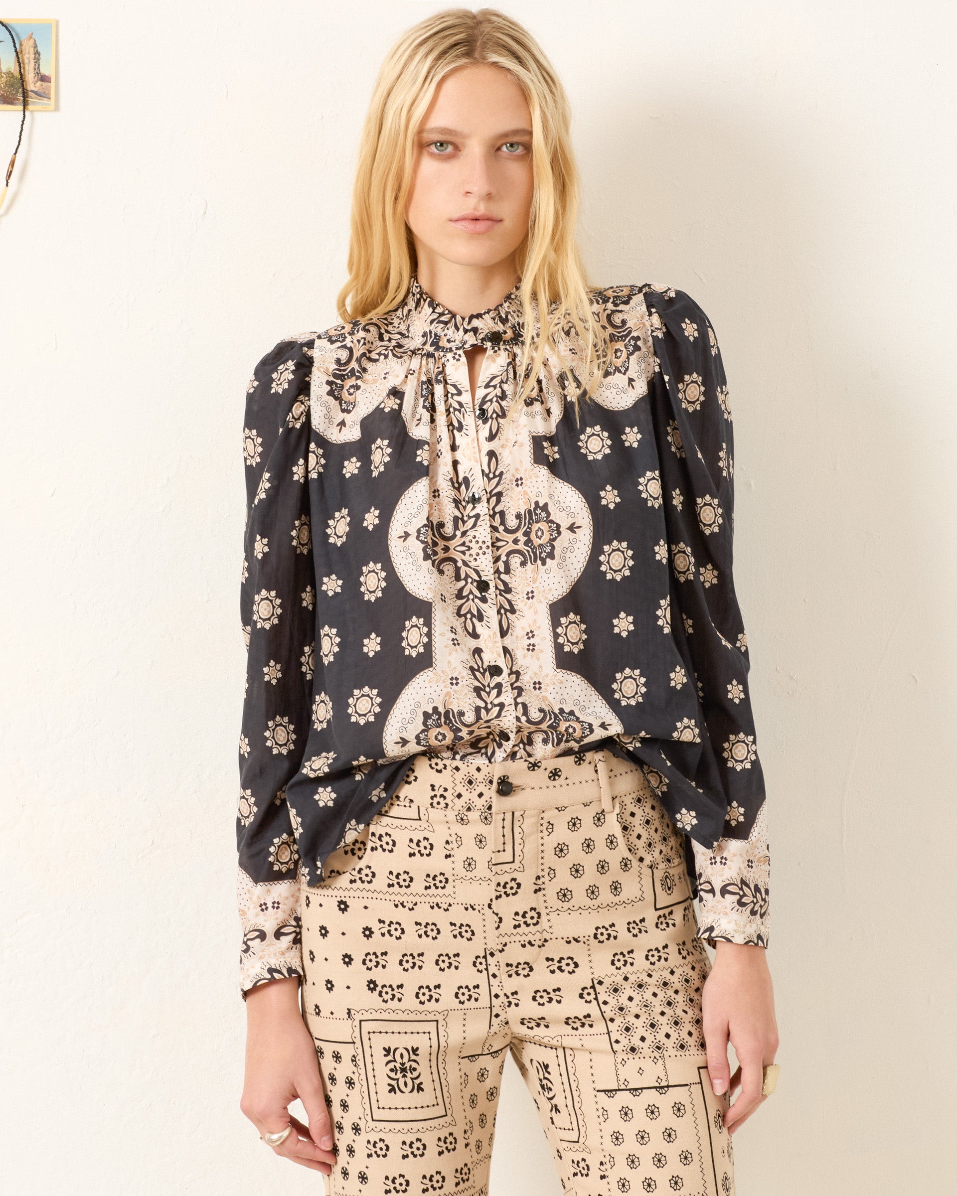 A woman with long blonde hair wears the Artemis Bronco Bandana Shirt, a cotton-silk blend with puff sleeves, paired with beige high-waisted pants featuring a black geometric print, standing against a plain light-colored wall.