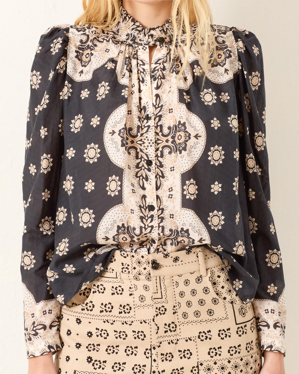 A woman wears the Artemis Bronco Bandana Shirt, a long-sleeved, cotton-silk blend top with a high neck and bow, paired with high-waisted, beige patterned pants.